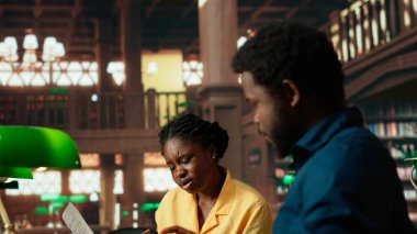 Siyahi üniversite öğrencileri üniversite kütüphanesinde sınıf notlarını karşılaştırıyor, akademisyenler fotokopi kitaplarından bilgi alıyor ve danışmanlık yapıyor. Öğrenciler yaklaşan sınavlarına hazırlanıyor. Kamera B.