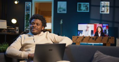 Neşeli Afrikalı Amerikalı adam şık bir oturma odasında dizüstü bilgisayarıyla telgraf çekiyor. Modern esnek çalışma alanında konfor ve üretkenliği dengeleyen uzaktan kumandalı bir işçi. Rahat bir çatı katı stüdyosu. Kamera B.