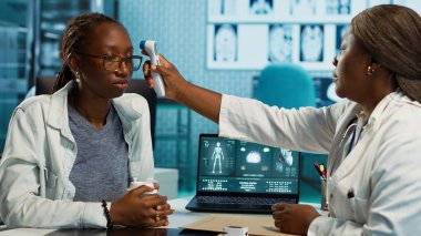 Klinikteki rutin kontroller sırasında sıcaklığı ölçen siyah sıhhiye grip belirtilerini kontrol etmek için dijital termometre kullanıyor. Sağlık uzmanı bir hastaya teşhis koymayı garanti ediyor. Kamera A.