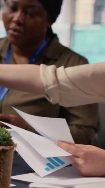 İş kadınlarından oluşan dikey video çeşitliliği ekibi, proje hedefleri belirlemek için şirket ofisindeki masanın etrafında toplanarak strateji raporlarını ve performans verilerini gözden geçiriyorlar. Ortaklık ve güçlenme. Kamera B.