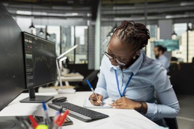 Malzeme firmasında çalışan tüccar, şamdan çizelgelerini ve fiyat grafiklerini incelerken not alıyor. İş yerinde çalışan Afrikalı Amerikalı kadın pazar eğilimleri ve ticaret stratejileri analizi yazıyor