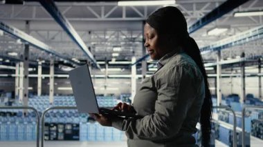 Bilgi işlem uzmanı tesiste yürürken dizüstü bilgisayarla sunucu çiftliği faaliyetlerini izliyor. Veri merkezindeki Afrikalı Amerikalı kadın sistemlerin düzgün çalıştığından emin olmak için test sonuçlarını gözden geçiriyor. Kamera A