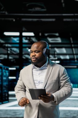 Veri merkezinde müzik dinleyen bir yönetici, altyapı ana bilgisayarlarını denetlemek için tablet kullanıyor. Sunucu çiftliğinde yürüyen, donanım donanımlarını denetlemek için tablet kullanan, ses kulaklığı takan işçiler