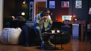 Genç siyahi adam oturma odasında serbest olarak çalışıyor, iş programını ayarlıyor ve rahat evinde çalışmaktan keyif alıyor. İnternette dizüstü bilgisayarla gezinen, birden fazla iş yapan biri. Kamera A.