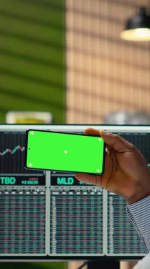 Ofisteki dikey video prop firması çalışanı yeşil ekran cep telefonundaki video dersinden yeni ticaret stratejileri öğreniyor. Afrikalı Amerikalı adam eğitim içeriğini izlemek için krom tuşlu akıllı telefon kullanıyor.