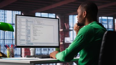 Ofisteki bilgisayarda kullanıcı arayüzüne bakan siyah bilişim uzmanı terminal penceresinde kod yazıp programlıyor. Çatı katında teknisyen tarafından kullanılan kodlama uygulaması. Kamera B.