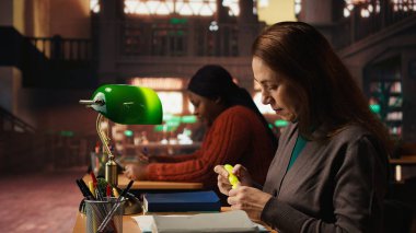 Kütüphanede önemli sınav fikirlerini vurgulayan, akademik materyal ve kitaplarla çevrili çalışmalarına odaklanan olgun bir kadın öğrenci. Strateji ezberlemesini çalış.