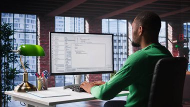 Ofisteki bilgisayarda kullanıcı arayüzüne bakan siyah bilişim uzmanı terminal penceresinde kod yazıp programlıyor. Çatı katında teknisyen tarafından kullanılan kodlama uygulaması. Kamera B.