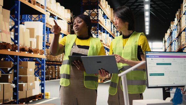 Team of diverse workers handling tracking software for parcel status on tablet, following the cargo shipment from the industrial warehouse with racks. Staff in hi vis vests. Camera A.
