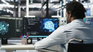 Paraplejik sunucu odası programcısı yapay zeka veri analizi grafiklerini yorumluyor. Tekerlekli sandalyedeki adam, veri merkezinde öngörü sistemi ölçümleri gösteren yapay zeka programı kullanıyor. Kamera B