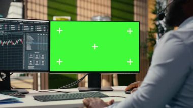 Yeşil ekran bilgisayar kullanan mali danışman müşteri aramaları sırasında yatırım rehberliği sunar. Kulaklık mikrofonuyla konuşan Afrikalı Amerikalı adam, müşterilere portföy yapımında yardım ediyor. Model PC, kamera B kullanarak.