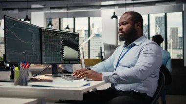 Bilgisayar masasındaki gülen portföy yöneticisi mal varlıklarını istatistiklere ve ekonomik göstergelere göre değerlendiriyor. Neşeli Afrikalı Amerikalı adam iş büyümesini yönlendirmek için araştırma raporlarına bakıyor, kamera A