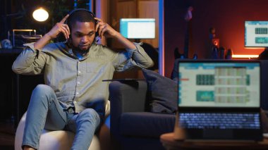 Siyah uzaktan kumandalı işçi ekrandaki analitik ekranın yanında geziniyor, evde oturuyor ve birden fazla cihazla günlük görevleri üzerinde çalışıyor. Çatı katında boş zaman ve iş verimliliği. Kamera B.