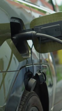 Elektrikli arabanın şarj cihazına bağlı dikey videosu sera gazı emisyonlarını azaltıyor, sürdürülebilir ulaşım kavramını azaltıyor. Kamu istasyonunda EV aracı şarj ediliyor, el kamerası görüntüsü