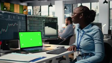 Finansal analist, yeşil ekran dizüstü bilgisayar kullanarak borsa hisseleri üzerinde araştırma yapıyor. Ofisteki Afrikalı Amerikalı işçiler takas kararları almak için maket defterindeki raporları okuyorlar, kamera A