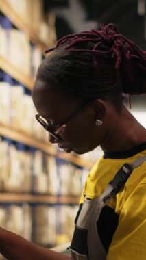 Dikey Afrikalı Amerikalı kadın ambardaki paketlere etiket yapıştırıyor, envanter ve sipariş işlemlerini yanlış etiketlerle yapıyor. Çevrimiçi alışveriş büyük iş deposu