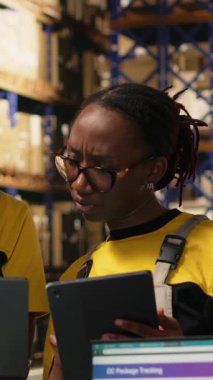 Dikey Video Deposu personeli paketleri denetliyor laptop ve tabletteki bilgileri takip ediyor, sevkiyata hazırlanıyor. Rafları, karton kutuları ve işlem aletleri olan organize bir depo. Kamera B.