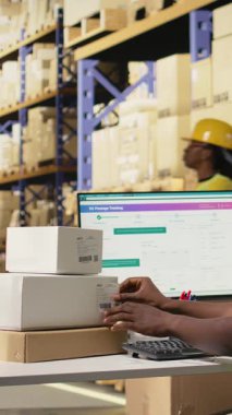 Dikey Afrikalı Amerikalı adam barkod işlemek için tarayıcı kullanıyor ve endüstriyel depolama raflarındaki kutulara nakliye etiketleri yapıştırıyor. Paket izleme servisi için yapıştırıcı etiketleri taranıyor