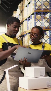 Lojistik merkezindeki sipariş işlemleri için dizüstü bilgisayardaki paket verilerini ele alan Dikey Video Siyah ekip üyeleri, dağıtım protokolleri için palet gönderme etiketlerini doğruladılar. Verimli bir e-ticaret deposu