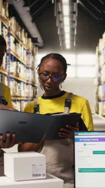 Dikey Video Deposu personeli paketleri denetliyor laptop ve tabletteki bilgileri takip ediyor, sevkiyata hazırlanıyor. Rafları, karton kutuları ve işlem aletleri olan organize bir depo. Kamera A.