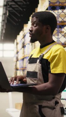 Dikey Video Deposu kâtibi büyük bir dağıtım merkezindeki raf ünitelerinin detaylarını inceliyor. Depoda güvenlik üniforması giyiyor. Malzeme zinciri yönetimi ve depolama odasına sevk