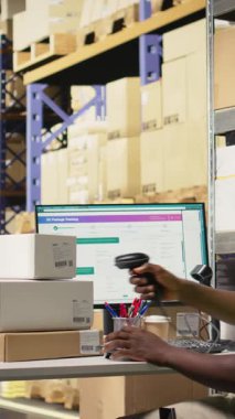 Dikey Afrikalı Amerikalı erkek çalışan, büyük bir depoda paketleri tarıyor, endüstriyel depo kayıtları içinde havayolu sevkiyat faturalarını kaydediyor. Sıralama tamamlamak için tarayıcı kullanılıyor