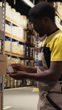 Dikey Video Afrikalı Amerikalı katip ambardaki ambalajlara etiket yapıştırıyor, envanter ve sipariş işlemleriyle uğraşıyor. Çevrimiçi alışveriş büyük iş deposu