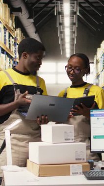 Dikey Video Siyah işçileri büyük bir depoda sevkiyat için mal hazırlıyor, envanterleri kontrol ediyor ve kargo dağıtımını yönetiyor. Lojistik siparişleri için paketleri sıralıyorum. Kamera A.