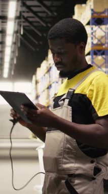 Dikey Video Afrikalı Amerikan e-ticaret işçisi nakliye etiketlerinin numaralarını tarıyor, envanter kayıtları için bavul ve sevkiyat detaylarını kaydediyor. Palet dağıtım işlemleriyle ilgilenen genç adam