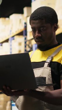 Dikey Video Genç zenci çalışanı büyük bir depo tesisinde raflarda paketleri ayırıyor. Paketlenmiş malların etrafındaki envanter ile birlikte yönetiliyor ve yerine getirilmesi için dağıtıma hazır.