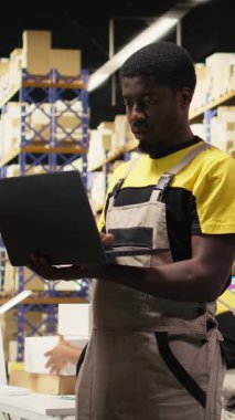 Dikey Video Genç zenci çalışanı büyük bir depo tesisinde raflarda paketleri ayırıyor. Paketlenmiş malların etrafındaki envanter ile birlikte yönetiliyor ve yerine getirilmesi için dağıtıma hazır.