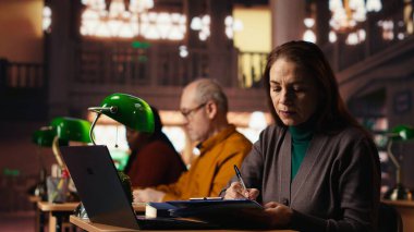 Entelektüel olgun bir kadın üniversitedeki dizüstü bilgisayarından akademik metni kopyalıyor, klasik çalışma odasında oturuyor ve çevrimiçi dokümanları tamamlıyor. Resmi öğrenim yoluyla eğitim.