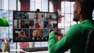 Masadaki Afrikalı Amerikalı adam iş arkadaşlarıyla görüntülü görüşme yapıyor, son iş haberleri hakkında konuşuyor. Uzaktan kumandalı işçi apartmandan sanal toplantıya katılıyor. Kamera A.