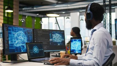 Ofisteki bilgisayar bilimcisi yapay zeka öğrenme sistemlerini iş akışlarını düzene sokacak şekilde entegre ediyor, kulaklık takıyor. Afrikalı Amerikalı adam cihazlar, dinleme müziği ve kamera A üzerinde yapay zeka kullanıyor.