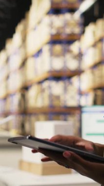 Dikey Video Siyah Kadın tabletteki depo lojistik sistemindeki teslimat kayıtlarını kontrol ediyor. Envanter ve ambalaj faaliyetleri raf birimleri içinde gerçekleşir, doğru sıranın yerine getirilmesini sağlar. Kamera.