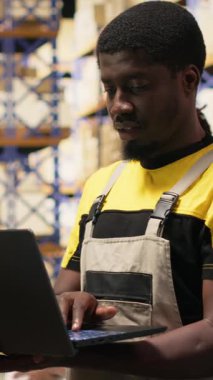 Dikey Video Siyah erkek personel e-ticaret lojistik merkezinde envanter paletleri doğruladı. Dizüstü bilgisayarda doğru işlem ve büyük ölçekli depo ayarlarında hızlı nakliyat için ambalaj