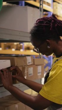 Dikey Afrikalı Amerikalı kadın ambardaki paketlere etiket yapıştırıyor, envanter ve sipariş işlemlerini yanlış etiketlerle yapıyor. Çevrimiçi alışveriş büyük iş deposu