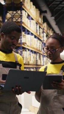 Dikey video Afrikalı Amerikan personeli envanter ve sevkiyat detaylarını kontrol ediyor. İş akışı ve verimli mal takibini sağlamak için büyük ölçekli dağıtım işlemleri