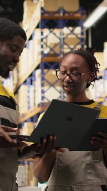 Dikey video Afrikalı Amerikan personeli envanter ve sevkiyat detaylarını kontrol ediyor. İş akışı ve verimli mal takibini sağlamak için büyük ölçekli dağıtım işlemleri