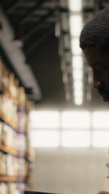 Dikey Afrikalı Amerikalı adam, tarayıcı kullanarak envanterdeki barkod etiketlerini kaydediyor, endüstriyel depodaki lojistik operasyonları çözüyor. İşçi, depolama raflarının yakınındaki nakliye etiketlerini kontrol ediyor