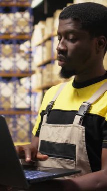 Dikey Video Afrikalı Amerikalı erkek personel, yoğun depolama merkezindeki ambalajları düzenliyor. Raflarda dizüstü bilgisayarla ve ürünlerle envanter tutuyordum. E-ticaret düzeni işleme