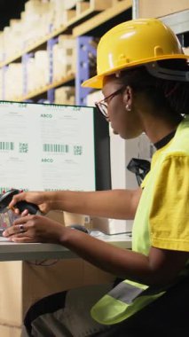 Dikey Video Siyah kadın, yelek ile tarayıcıyla takip bilgilerini kaydediyor, kargo siparişleri için ekspres teslimat servisini denetliyor. Etiketler üzerindeki sevkiyat detayları taranıyor, pc yazılımı izleniyor