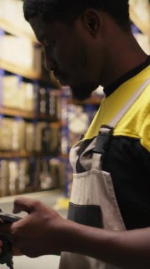 Dikey Afrikalı Amerikalı adam, tarayıcı kullanarak envanterdeki barkod etiketlerini kaydediyor, endüstriyel depodaki lojistik operasyonları çözüyor. İşçi, depolama raflarının yakınındaki nakliye etiketlerini kontrol ediyor