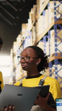 Dikey Video Siyah işçileri büyük bir depoda sevkiyat için mal hazırlıyor, envanterleri kontrol ediyor ve kargo dağıtımını yönetiyor. Lojistik siparişleri için paketleri sıralıyorum. Kamera B.