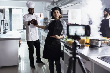 Önlüklü profesyonel kadın aşçı bir restoranda yemek pişirme dersi kaydediyor, tekniklerini ve bilgilerini çevrimiçi seyircilere paylaşıyor. Kadın yemek hazırlıklarının her adımını kaydediyor..