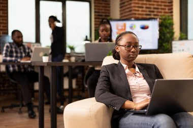 İşletme analizlerini gözden geçiren, odaklanmış ifadesiyle dizüstü bilgisayarda çalışan Afrikalı Amerikalı kadın girişimci. Gözlüklü profesyonel bir kadın, rahat bir koltukta oturuyor ve kişisel bilgisayar kullanıyor..