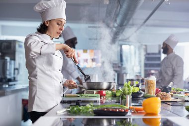Üniformalı kadın profesyonel şef tavada bir gurme yemeği hazırlıyor, sıcak bir sobanın üzerinde taze sebzeler karıştırıyor. Lezzetli malzemeler güzel bir yemek şaheserine karıştığında buhar yükselir..
