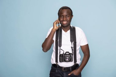 Gülümseyen siyah adam cep telefonundaki bir telefona cevap verirken bir eli cebinde kameraya bakıyor. Pantolon askısı takan erkek fotoğrafçı stüdyo çekimi için kulağına bir telefonla poz veriyor..