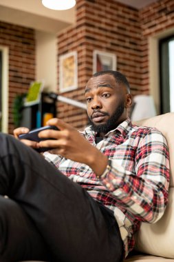 Uzaktan kumandalı erkek çalışan, tuğla duvarlı oturma odasında kanepede oturuyor, teneffüste cep telefonuyla oyun oynuyor. Afrikalı Amerikalı serbest oyuncu, mobil oyun sırasında rakibinin yaptığı hamleye şaşırdı..
