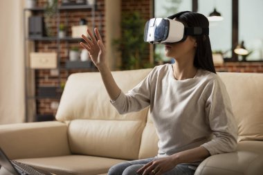 Odaklanmış iş kadını el kaldırılmış bir şekilde kanepeye oturmuş, duvardaki evde sanal gerçeklik kulaklığı takıyor. Serbest çalışan bir kadın, teknolojiyi araştırıyor, iş haritalarını araştırıyor ve projeler planlıyor..